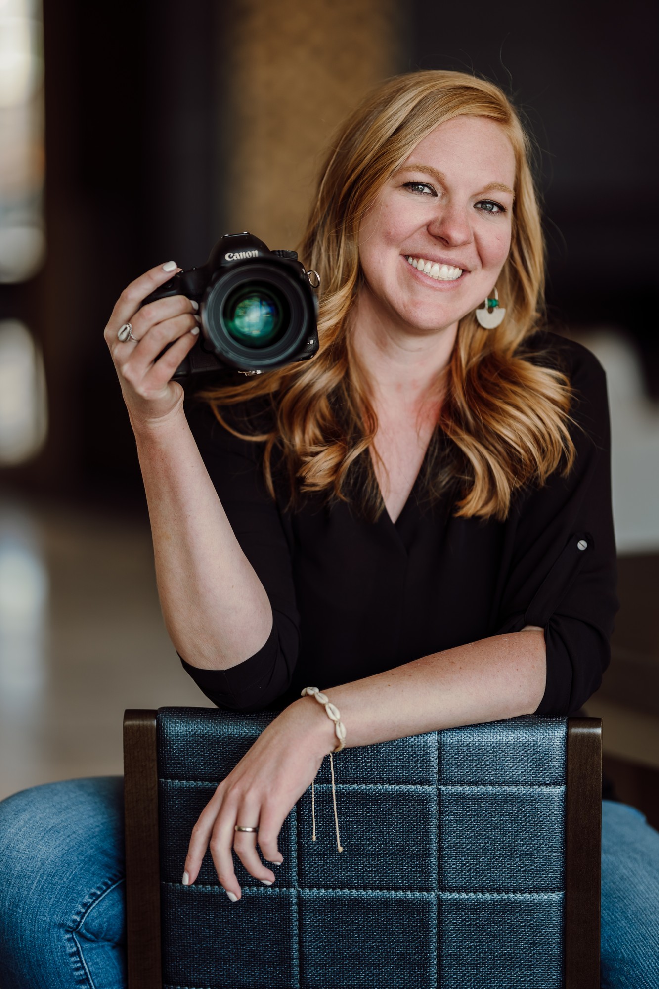 Rachel smiling and holding her Canon camera, arms resting on the back of a chair