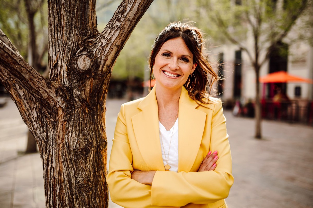 Client with a brunette ponytail in a yellow blazer, arms crossed beside a tree