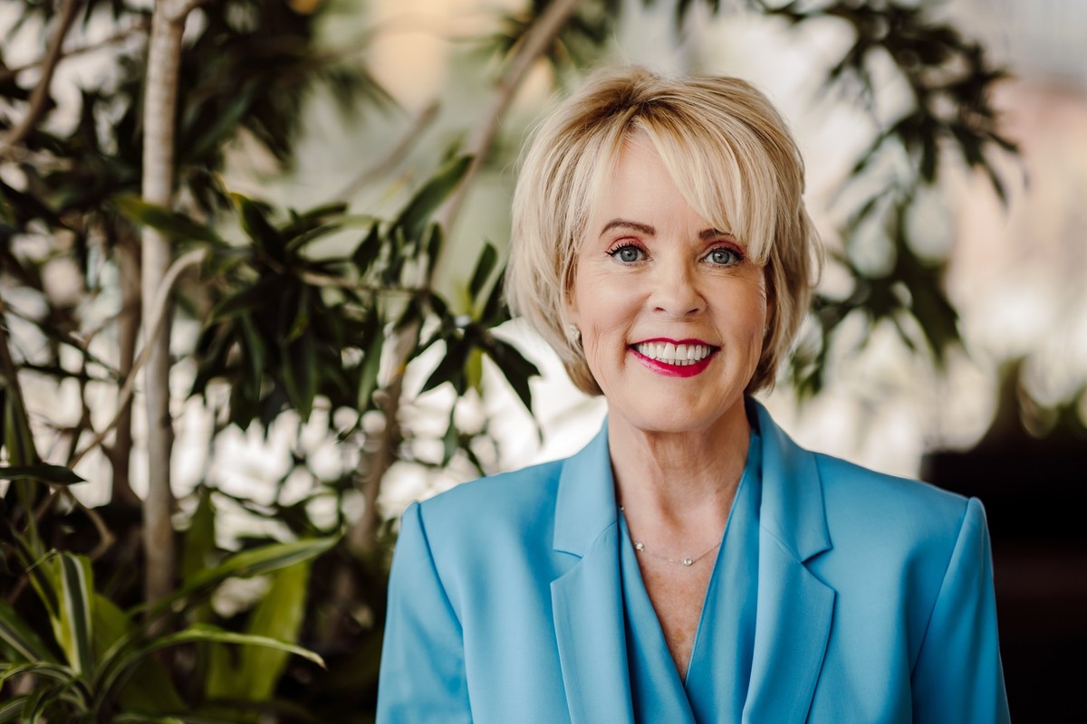 Client with a short blonde bob and red lipstick in a light blue suit, in front of leafy plants
