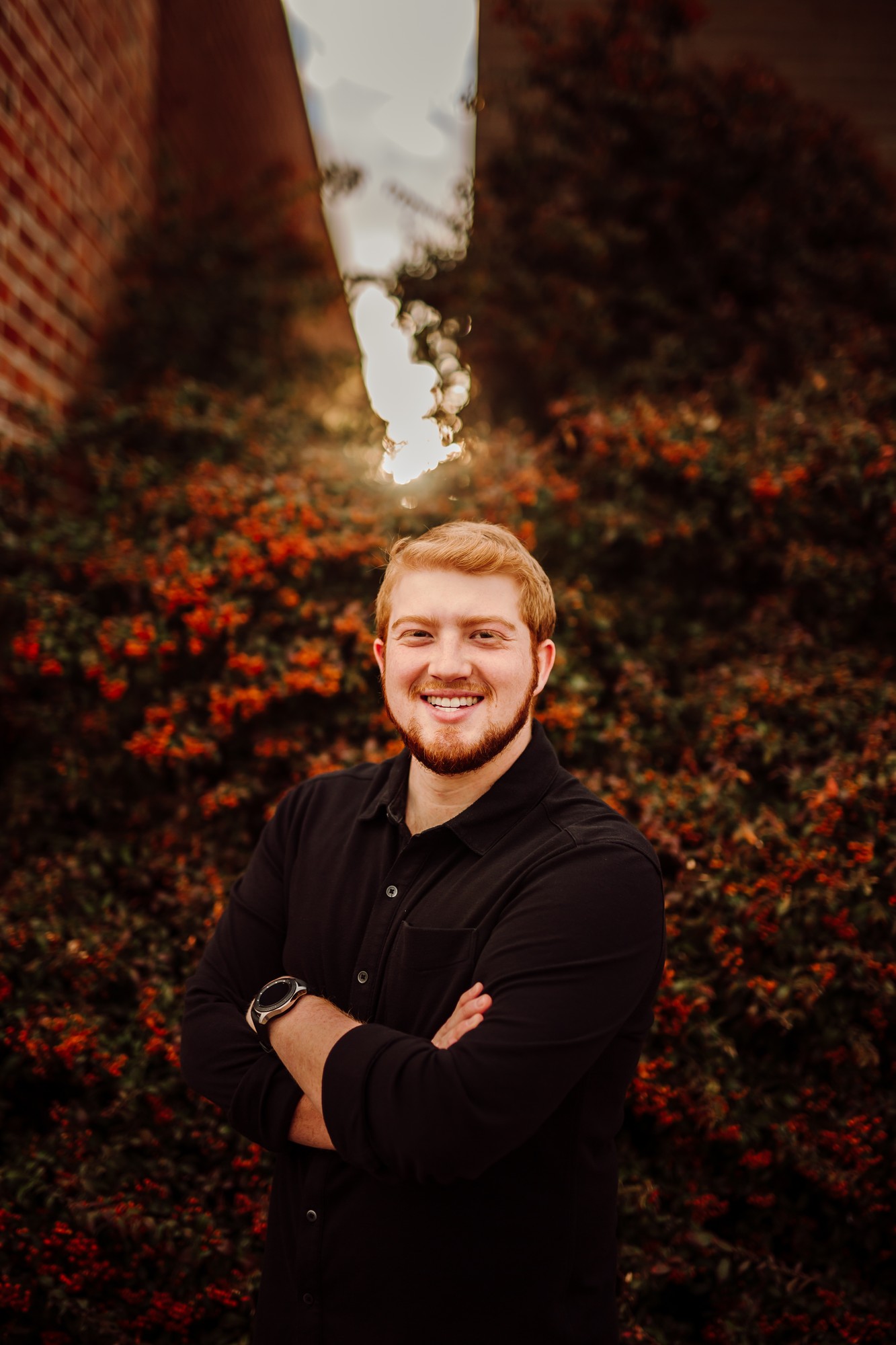 Headshot client with red hair and a beard in a black button-down, arms crossed in front of sunlit autumn shrubs