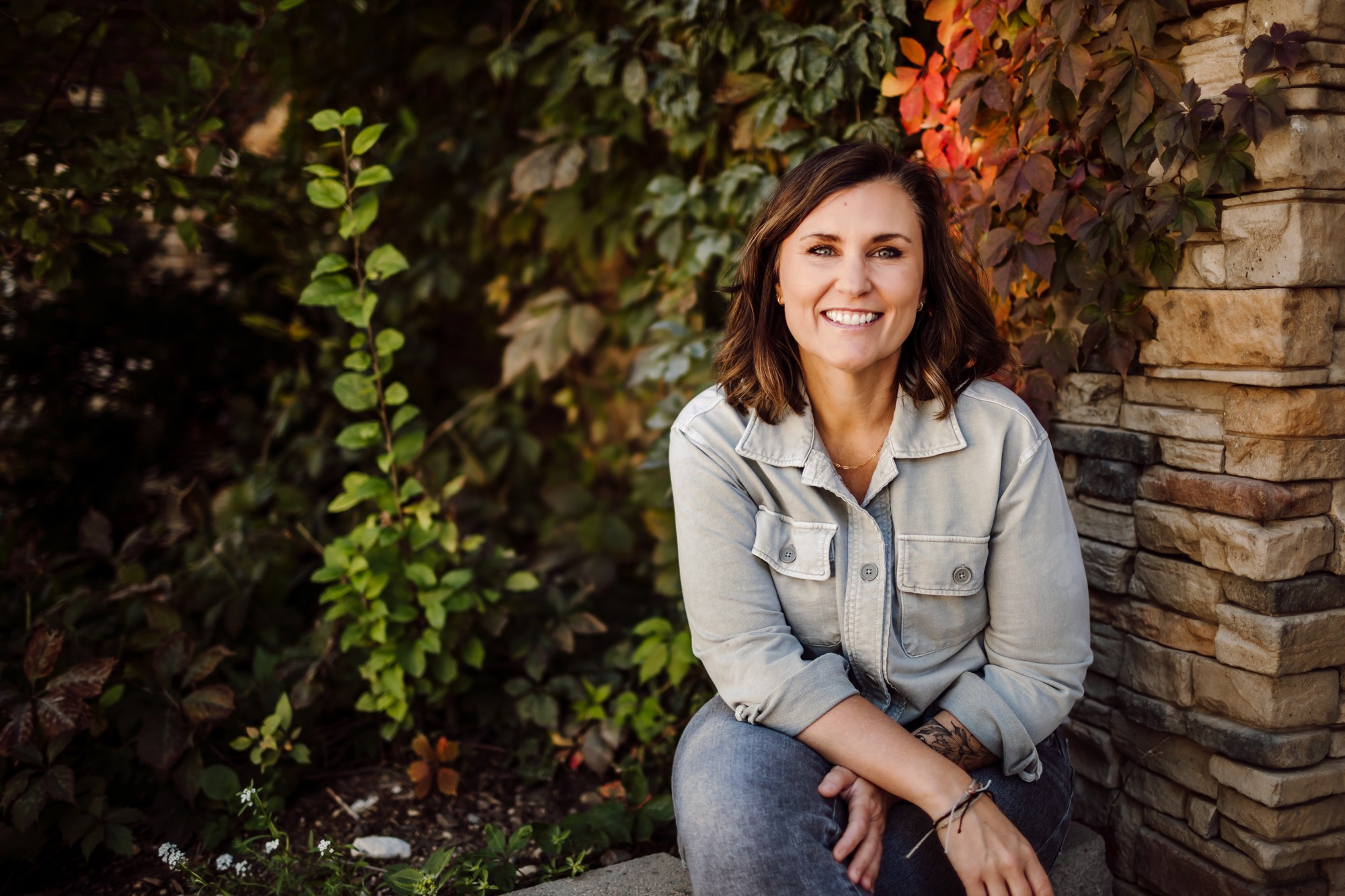 Headshot client in a light denim shirt seated beside a stone pillar wrapped in autumn vines