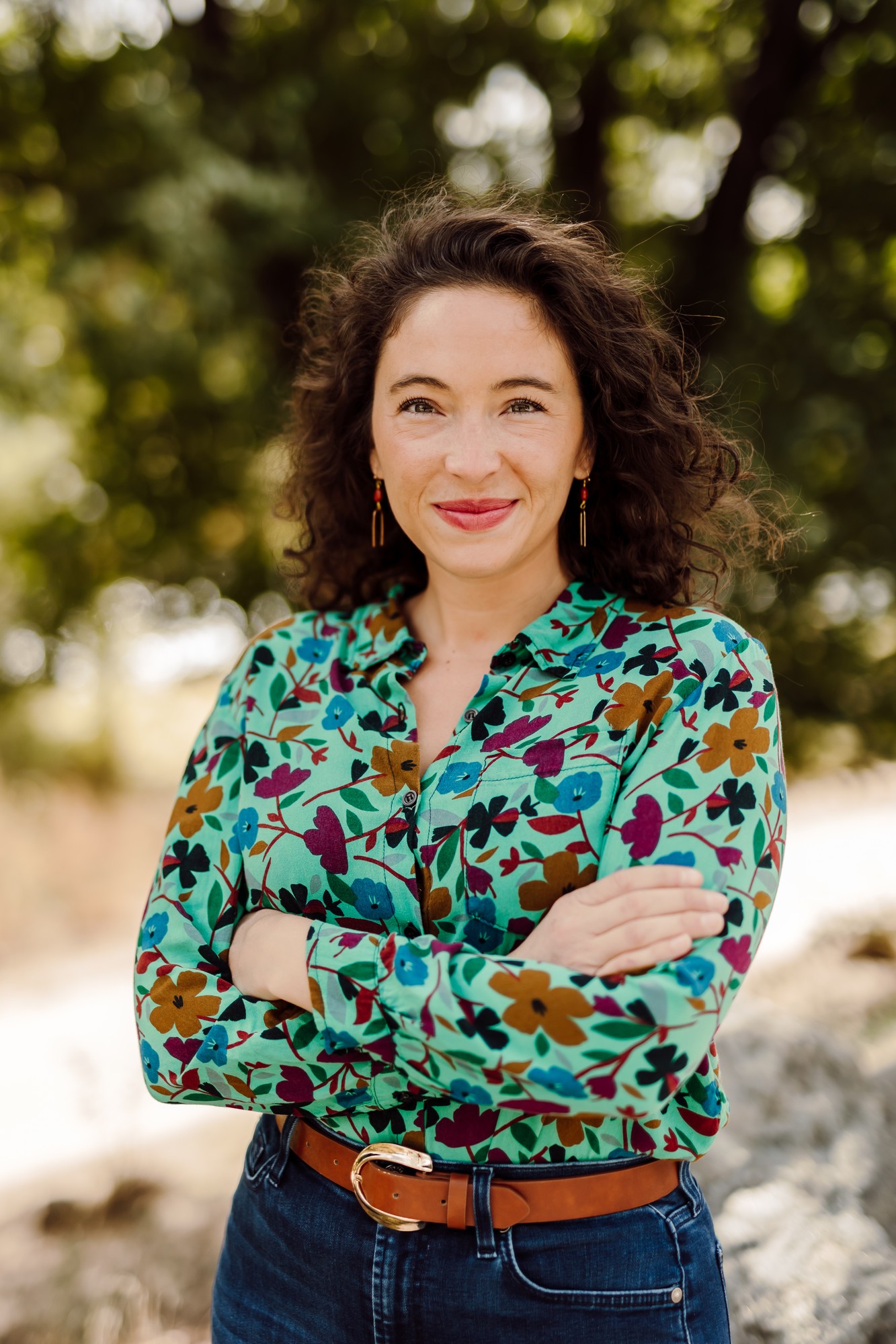 Headshot client with curly hair in a green floral shirt, arms crossed beneath the trees