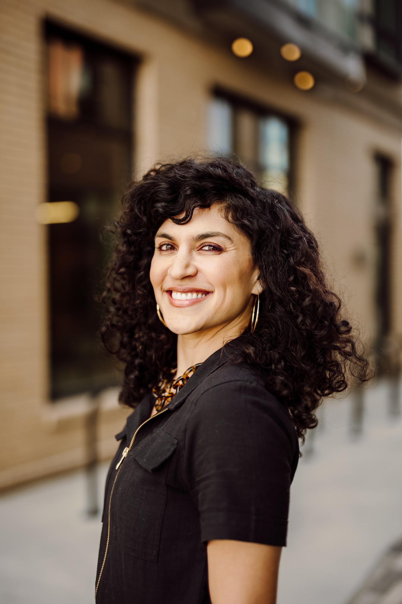 Headshot client with dark curls in a black shirt smiling over her shoulder downtown
